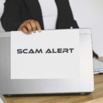 Pracownik trzyma tabliczkę scam alert nad laptopem w biurze